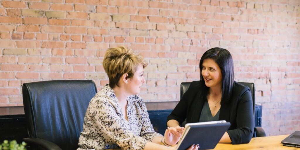 Two women sit at a wooden table, engaged in discussion. One holds a tablet. They appear professional and focused, set against a brick wall backdrop.
