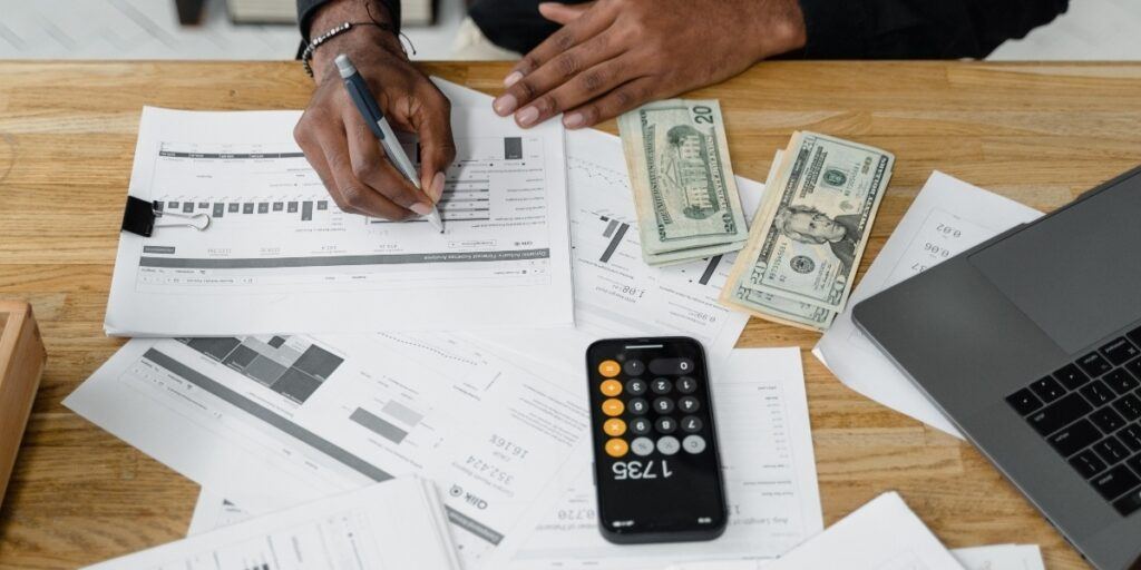 A person works at a wooden table covered with financial documents, using a calculator and pen. Cash and a laptop are also visible, evoking productivity.
