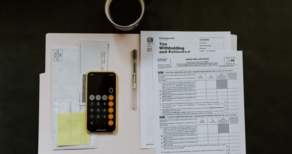 A desk with tax forms, a calculator app on a smartphone, a pen, a sealed envelope, and a cup of coffee. The scene suggests financial planning.