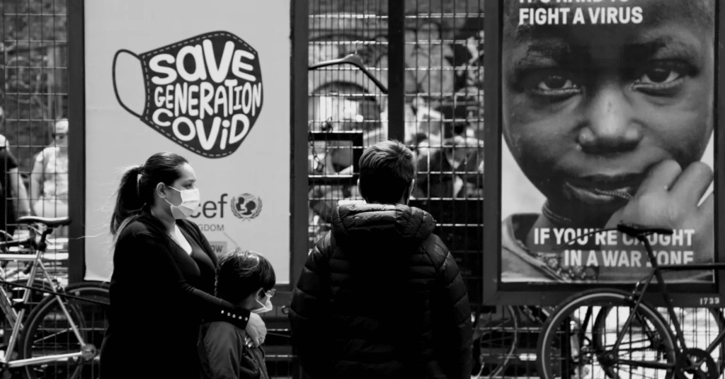 A black-and-white photo shows a woman and two children wearing masks, standing near posters. One reads "Save Generation Covid," another features a child's face urging to "fight a virus."