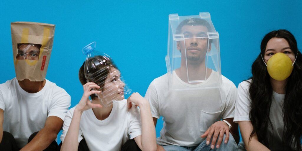 Four people in white shirts sit against a blue background, wearing makeshift masks: a paper bag, plastic bottle, container, and round yellow mask. Playful mood.