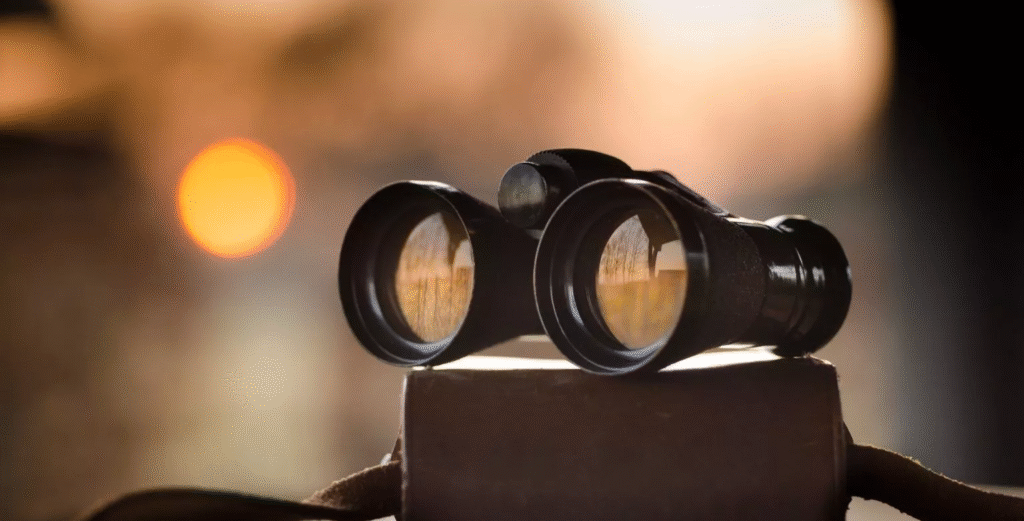Binoculars resting on a surface with soft focus background featuring a warm, glowing sunset. The mood is serene and contemplative.