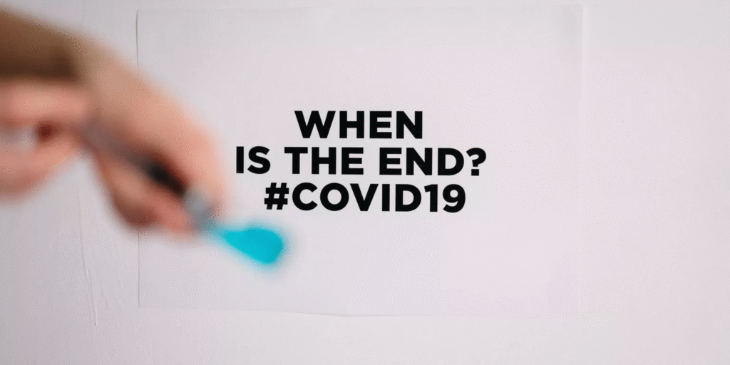 A blurred hand holding a blue paintbrush appears in front of a sign on a white wall that reads "WHEN IS THE END? #COVID19," conveying uncertainty and concern.