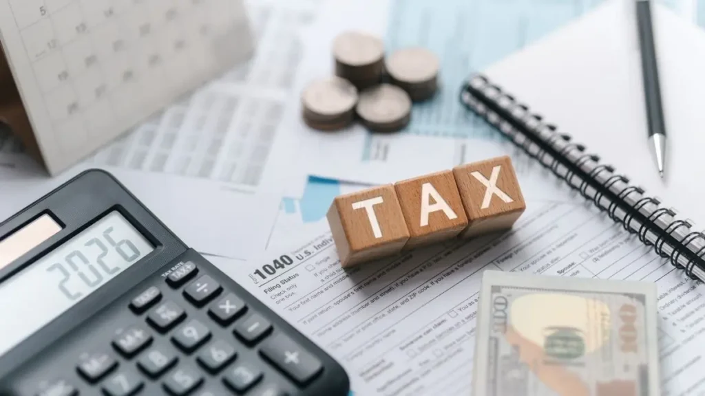 A calculator displaying "2023," wooden blocks spelling "TAX," coins, a spiral notebook, and dollar bills on tax forms, suggest financial planning.